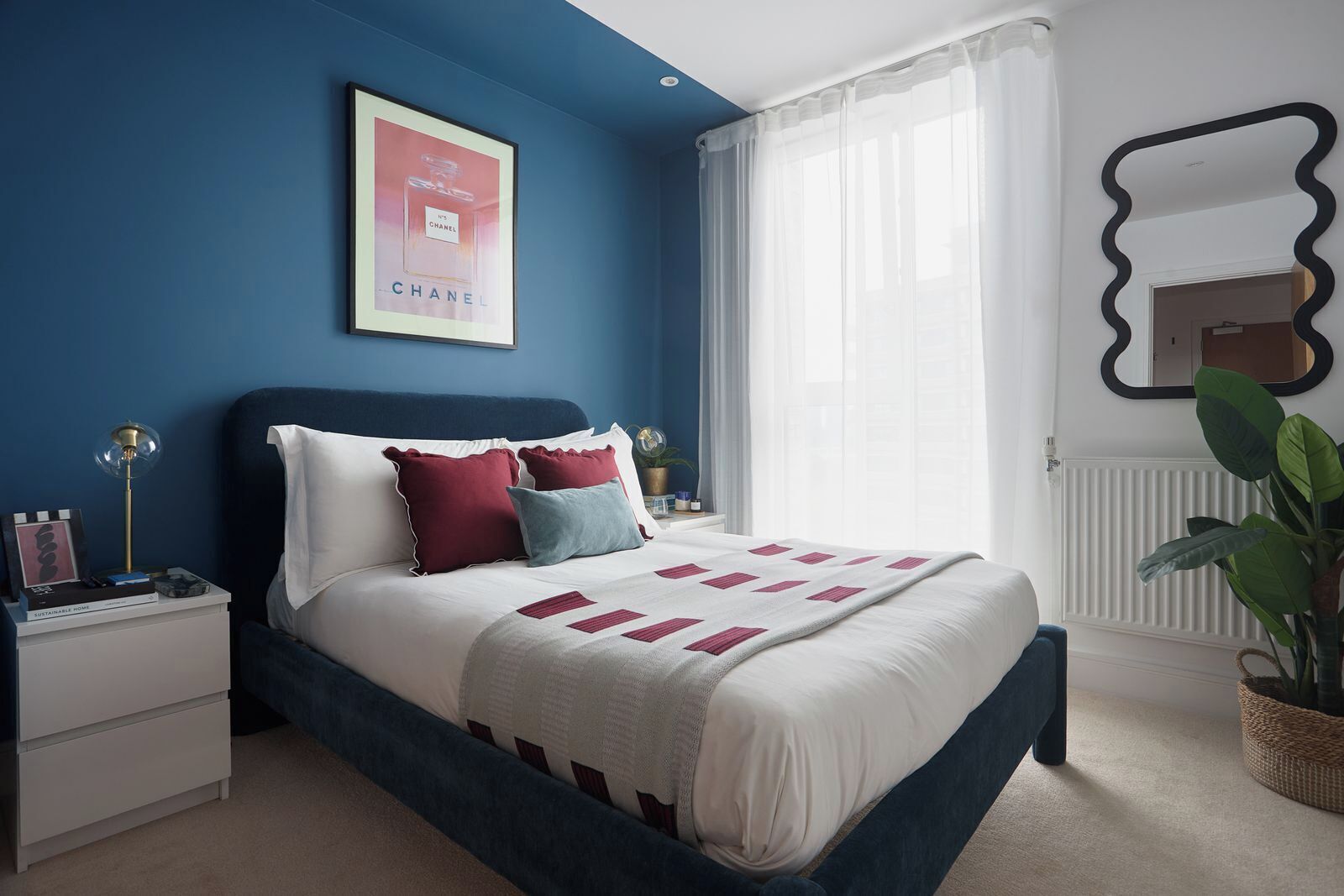 A bedroom with a bed, nightstands, a framed picture, a window with sheer curtains, and a mirror on the wall.