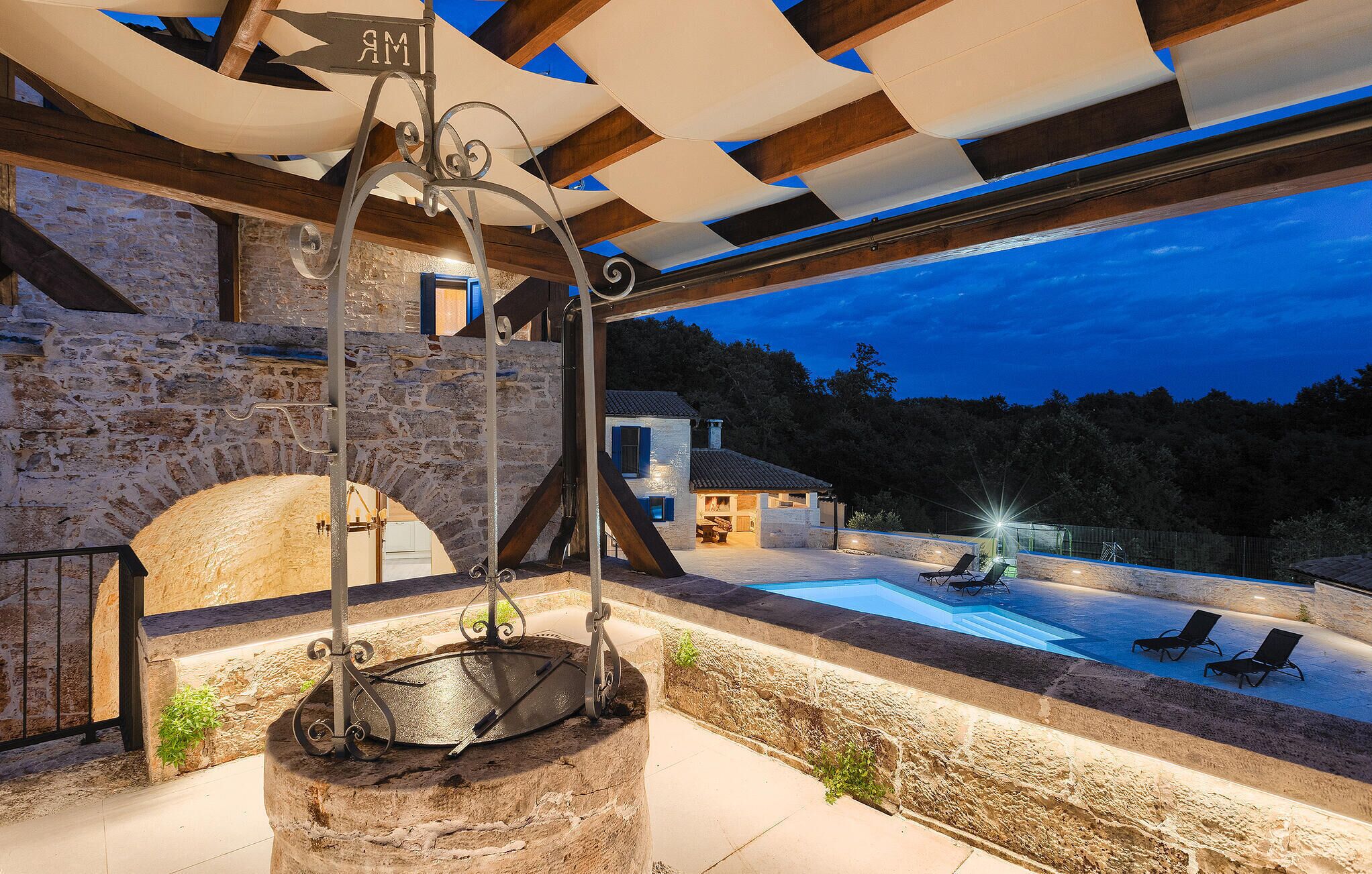 A stone structure with a metal spiral staircase, a pool area with lounge chairs, and a view of a house in the background.