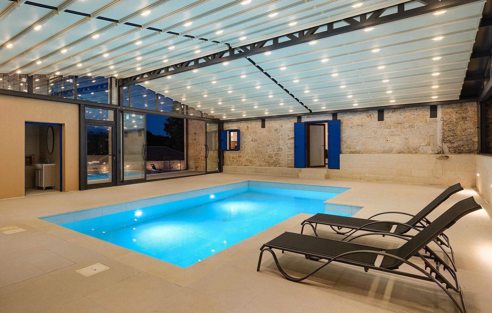 An indoor swimming pool with glass walls, a stone wall, and a covered area with a ceiling of lights.