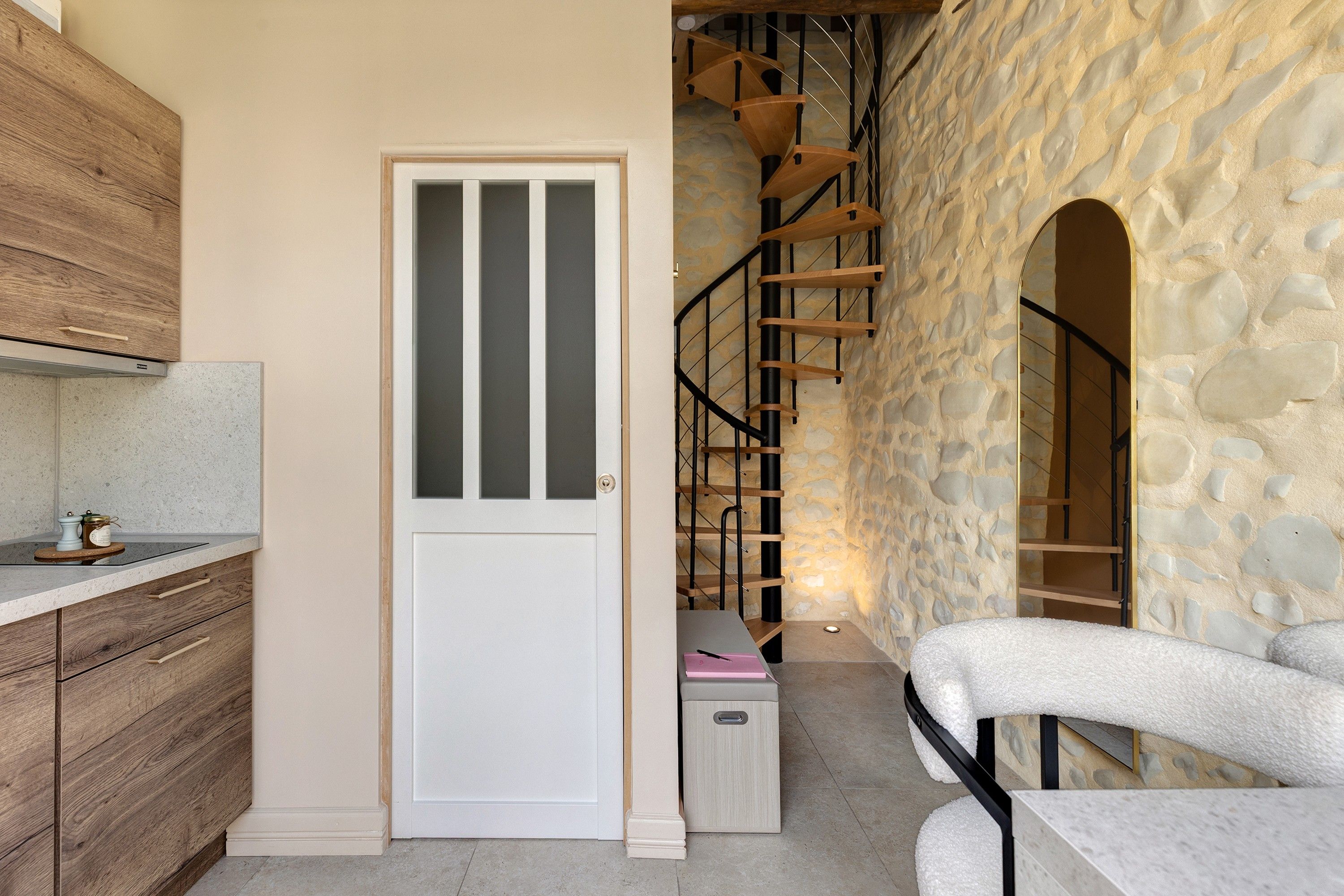 A kitchen with wooden cabinets, a white door with a frosted panel, a staircase with wooden steps, and a stone wall.