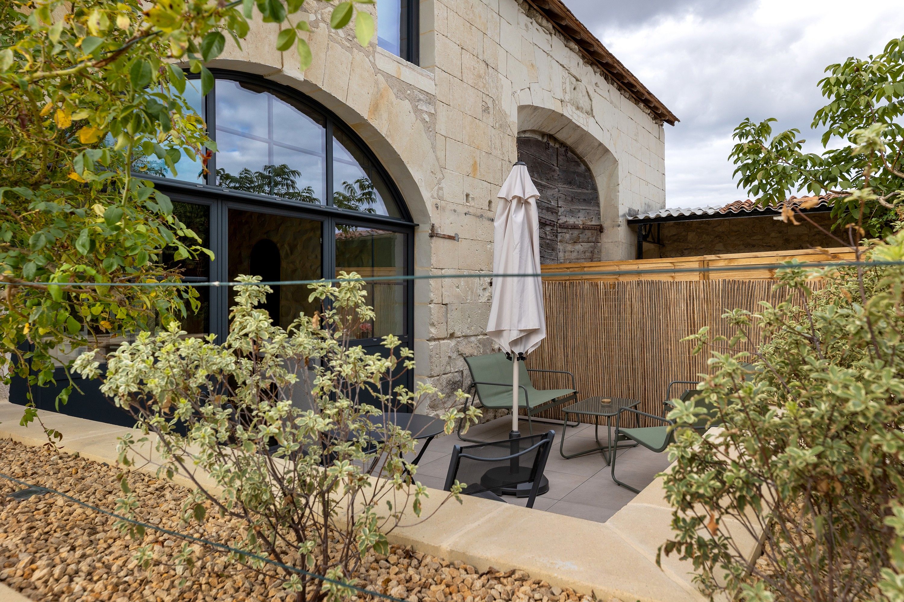Un bâtiment en pierre doté d’une grande fenêtre en arc de cercle, d’un patio couvert avec une table et des chaises, et d’une clôture en bambou.