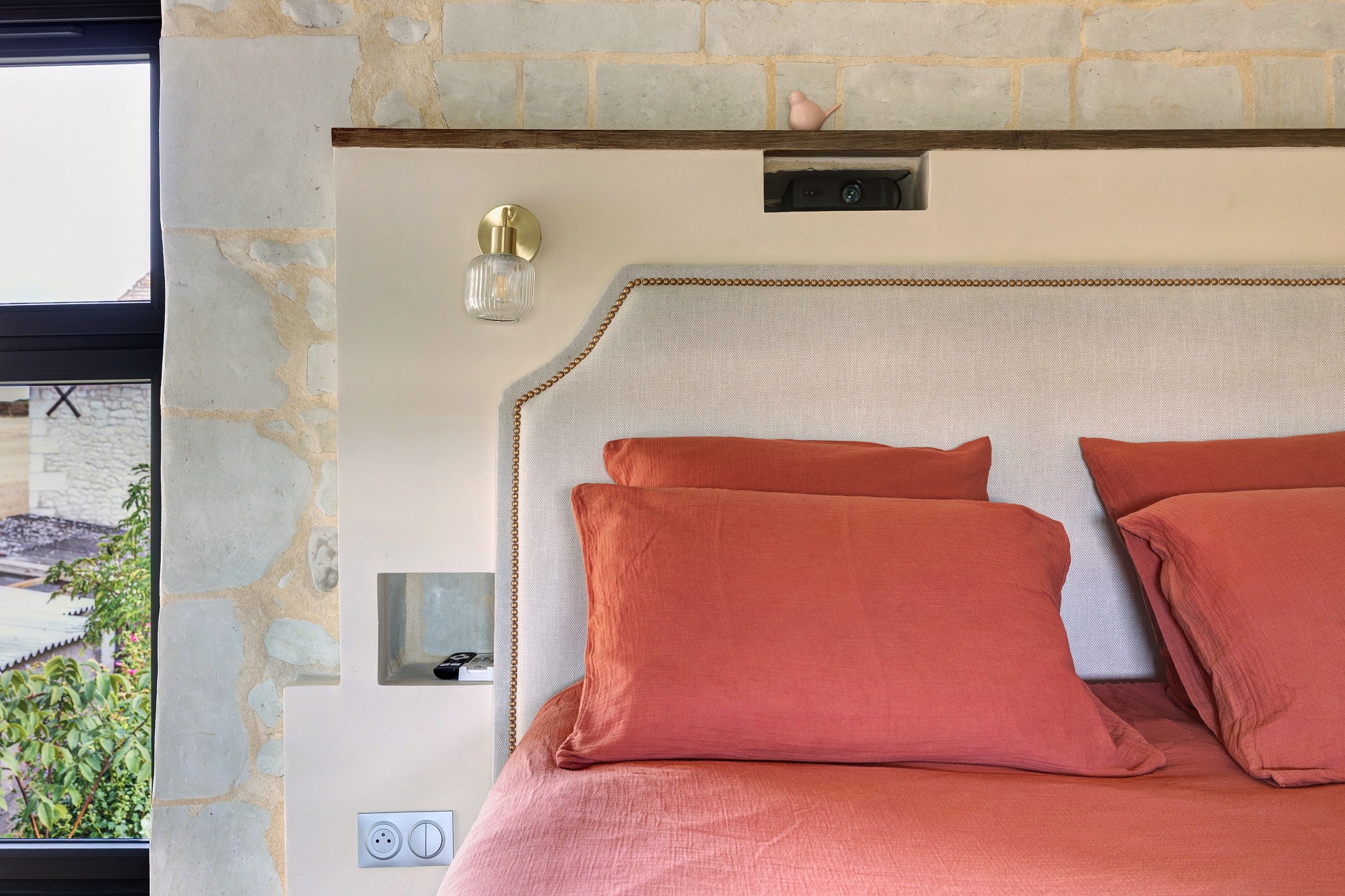Une chambre avec un mur en pierre, un lit avec deux oreillers, un luminaire fixé au mur et une petite étagère sur laquelle se trouve un appareil photo.