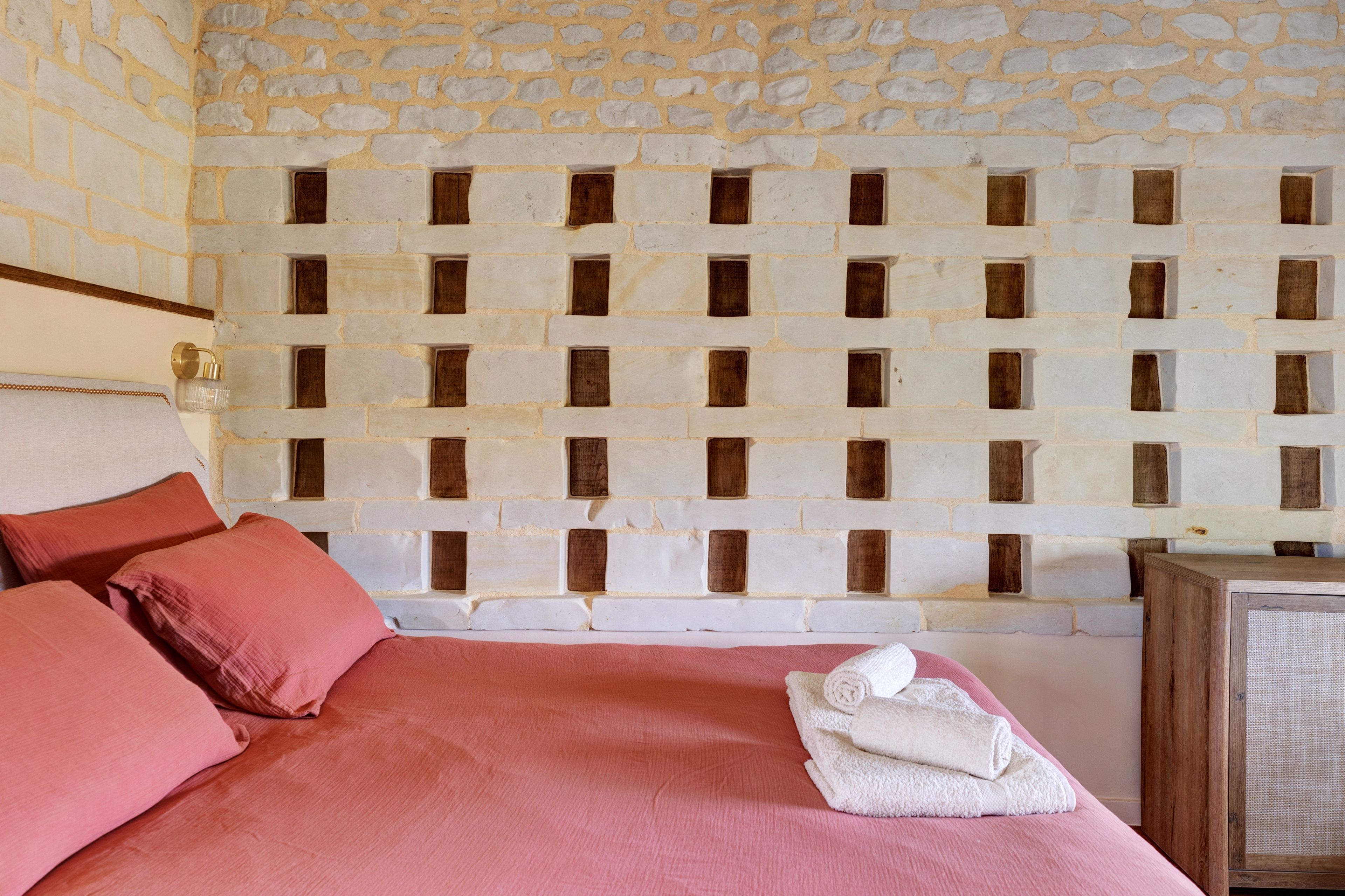 Une pièce comprenant un lit, des oreillers, un mur avec un motif en briques et une petite armoire en bois.