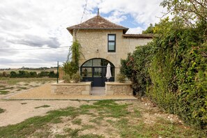 A stone building with a tiled roof, a large arched window, and a small patio area with a table and chairs. - Holiday Home “Le Pigeonnier Des Châpeaudieres” with Private Terrace, Wi-Fi, Air Conditioning (Châtellerault)