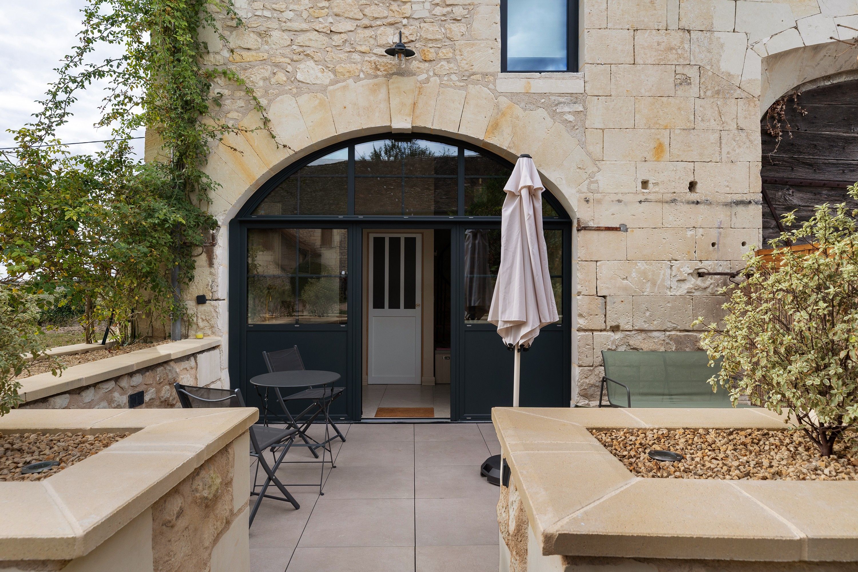 A stone building with a patio area featuring a table and chairs, a covered seating area, and a view through a large window.