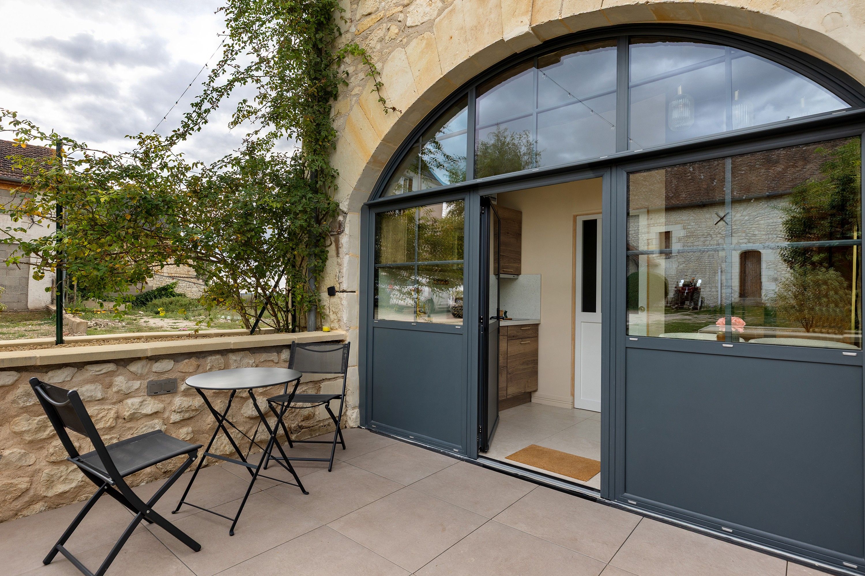 Une terrasse extérieure avec un mur en pierre, une table, deux chaises et une grande porte vitrée donnant sur une pièce comprenant une cuisine et offrant une vue sur un bâtiment en pierre.