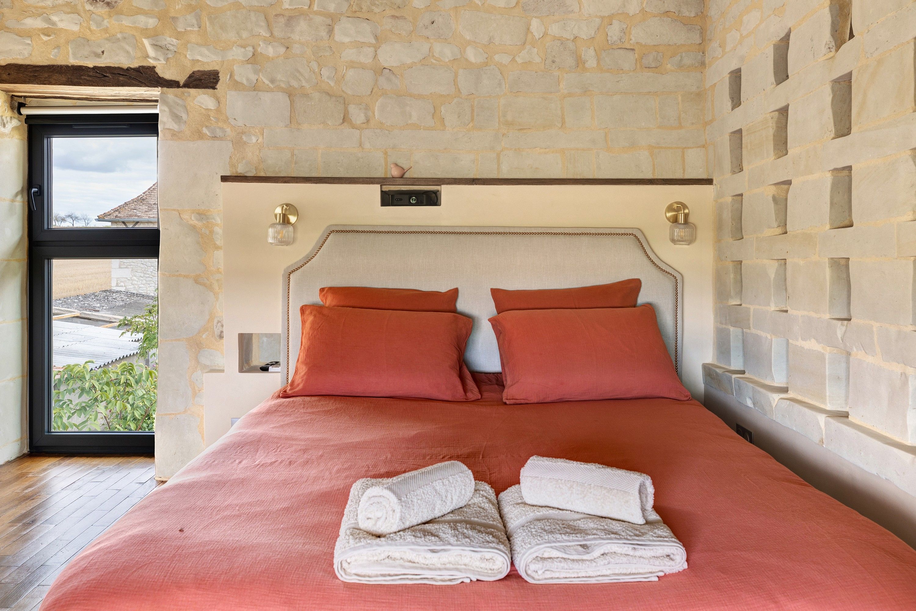 Un dormitorio con pared de piedra, una cama con dos almohadas y una ventana con vistas a edificios.