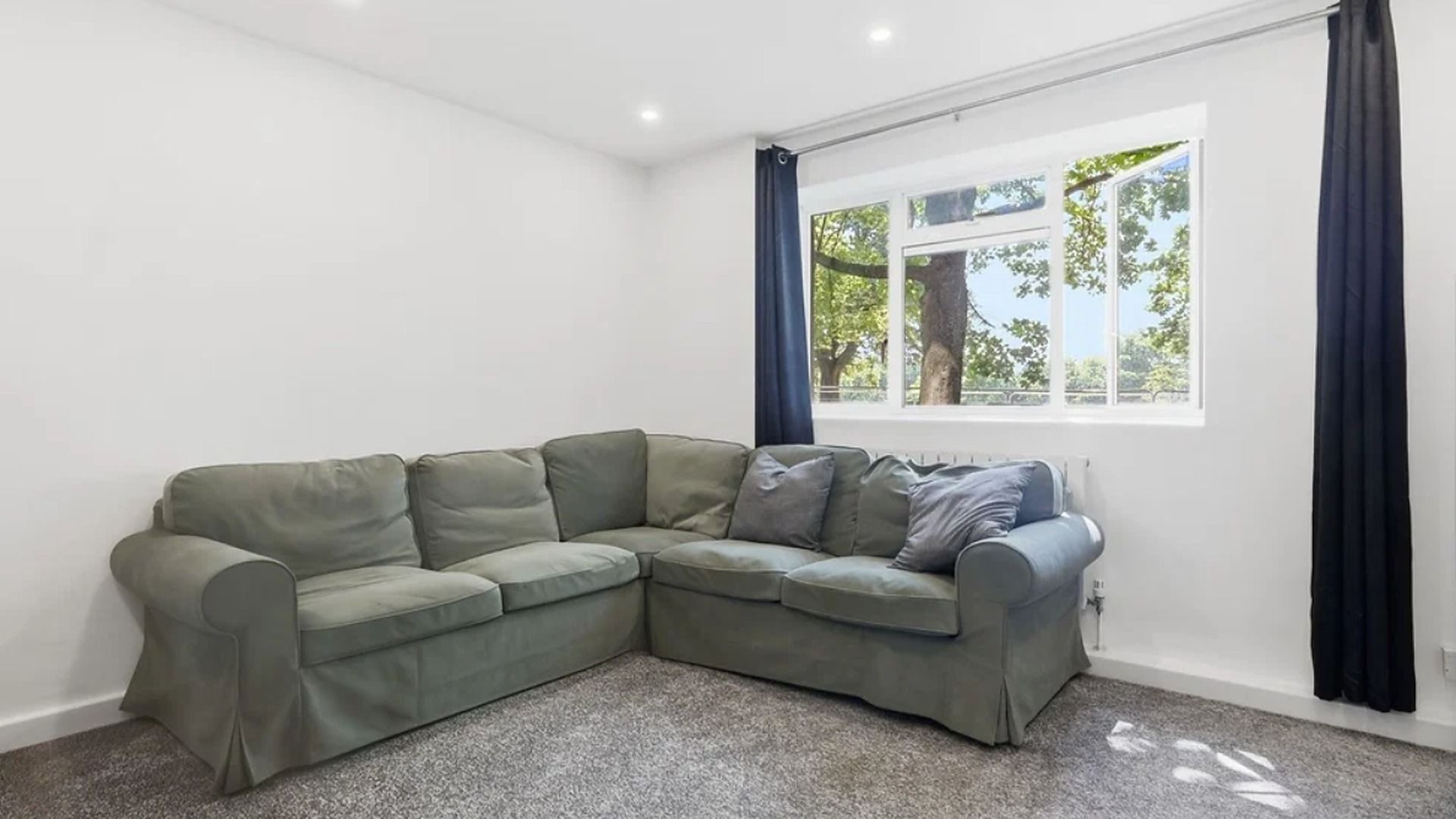 A corner living room with a sectional sofa, a large window with curtains, and a radiator.