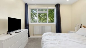 Een slaapkamer met een bed, een dressoir, een televisie, een raam met gordijnen en een radiator.