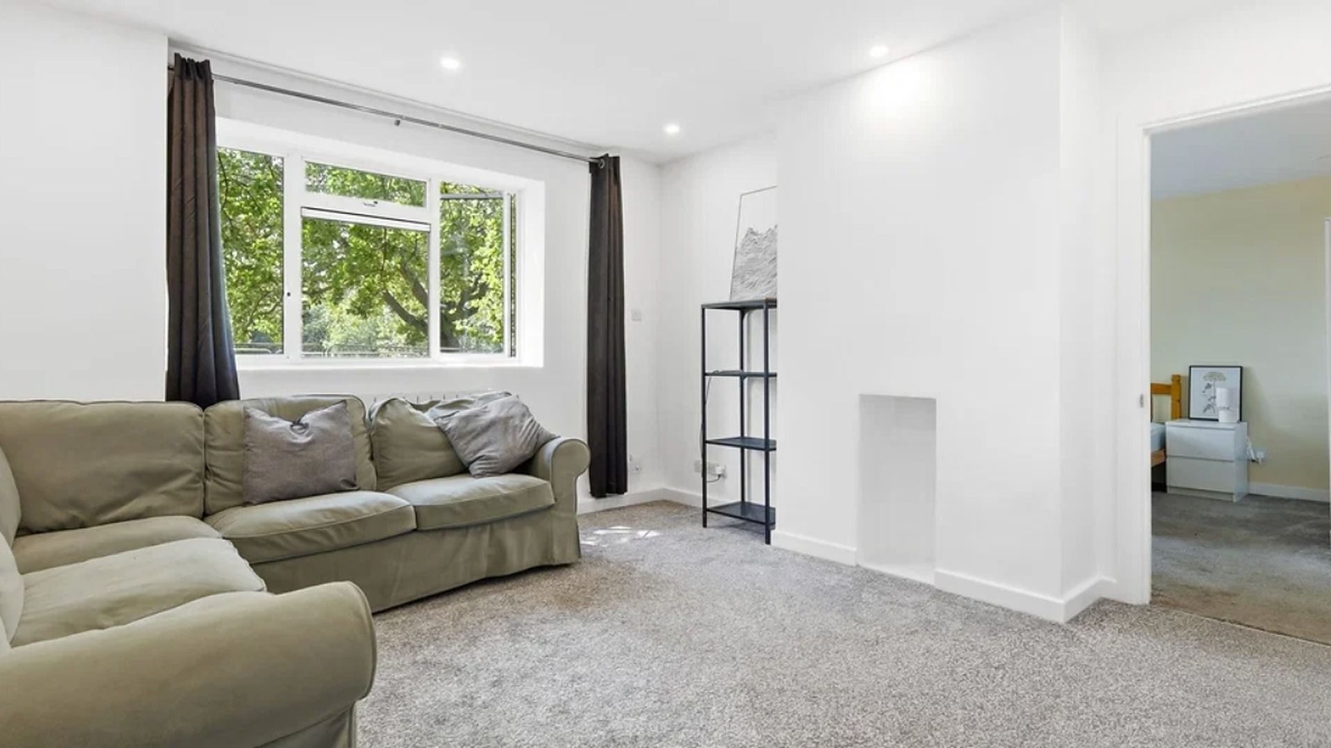 Et værelse med en beige sofa, en sort reol og et vindue med gardiner.
