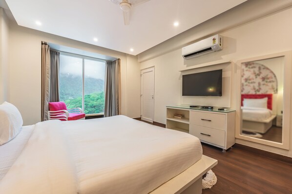 A hotel room with a large bed, a desk with a flat-screen TV, a wardrobe, and a sliding door leading to a balcony with a view of greenery.