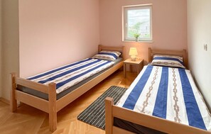 Ein Zimmer mit zwei Holzbetten, jeweils mit gestreifter Bettwäsche. Ein Fenster mit Jalousien, ein kleiner Tisch mit Lampe und ein Blumentopf auf der Fensterbank.