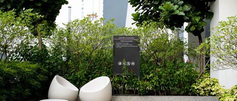 Un área de estar al aire libre con sillas blancas modernas, rodeada de abundante vegetación y un cartel que detalla las normas para una terraza infantil/de yoga.
