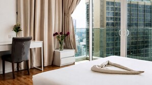 A hotel room with a bed, a desk, a chair, a vase with flowers, and a view of a cityscape through large windows. - Quill Residences KLCC by Moonlight (Kuala Lumpur)
