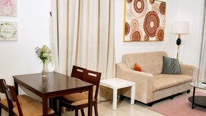 A living room with a beige sofa, a small table, a dining table with chairs, and a vase with flowers. - Quill Residences KLCC by Moonlight (Kuala Lumpur)