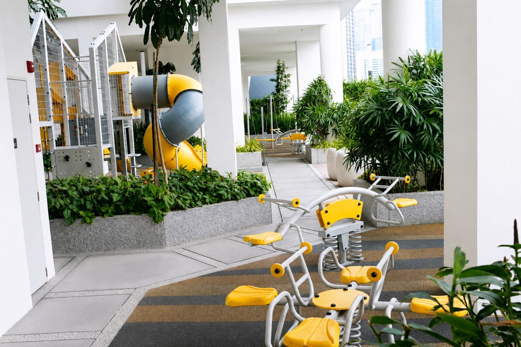 Un espacio exterior moderno con equipamiento de ejercicio, un tobogán amarillo y abundante vegetación.
