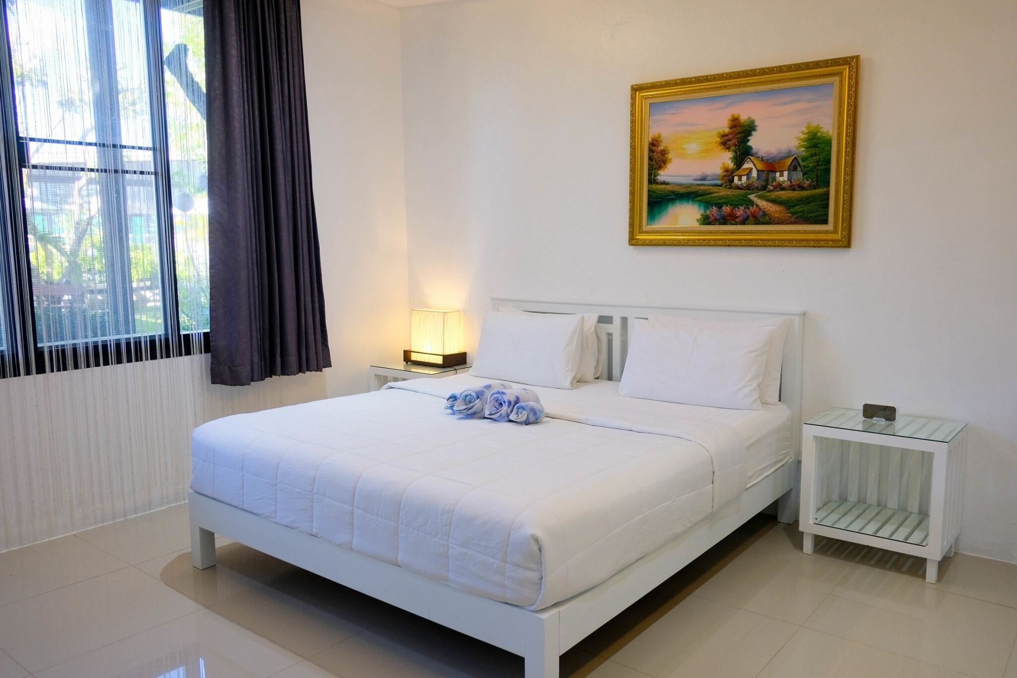 Quarto de hotel com uma cama grande, uma mesa de cabeceira com abajur, um quadro na parede e uma janela com cortinas.