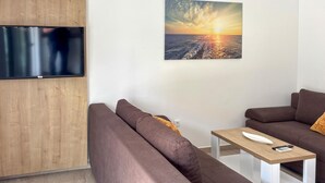 Ein modernes Wohnzimmer mit einem an einer Holzplatte befestigten Flachbildfernseher, zwei braunen Sofas, einem kleinen Holztisch mit einer Fernbedienung und einem Wandbild, das einen Sonnenuntergang über Wasser zeigt.