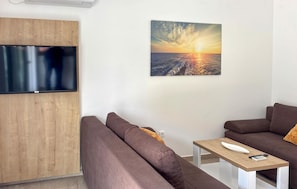 Ein modernes Wohnzimmer mit einem an einer Holzplatte befestigten Flachbildfernseher, zwei braunen Sofas, einem kleinen Holztisch mit einer Fernbedienung und einem Wandbild, das einen Sonnenuntergang über Wasser zeigt.