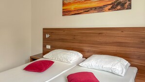 Ein Doppelbett mit weißen Bettwäsche und zwei roten Kissen, ein Holz-Kopfteil, ein Nachttisch mit Lampe sowie ein Sonnenuntergangsbild an der Wand.
