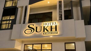En hotellbyggnad med flera våningar, med en tydlig skylt som visar namnet ”Hotel Sukh” och som tillhör Mehar Group.