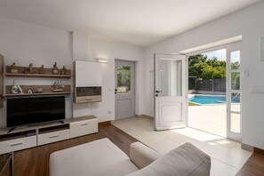 A modern living room with a flat-screen TV, wooden shelves, a white sofa, and a sliding glass door leading to a pool area. - Villa Pajari (100581-K1) - Sveti Lovrec (Sveti Lovrec)