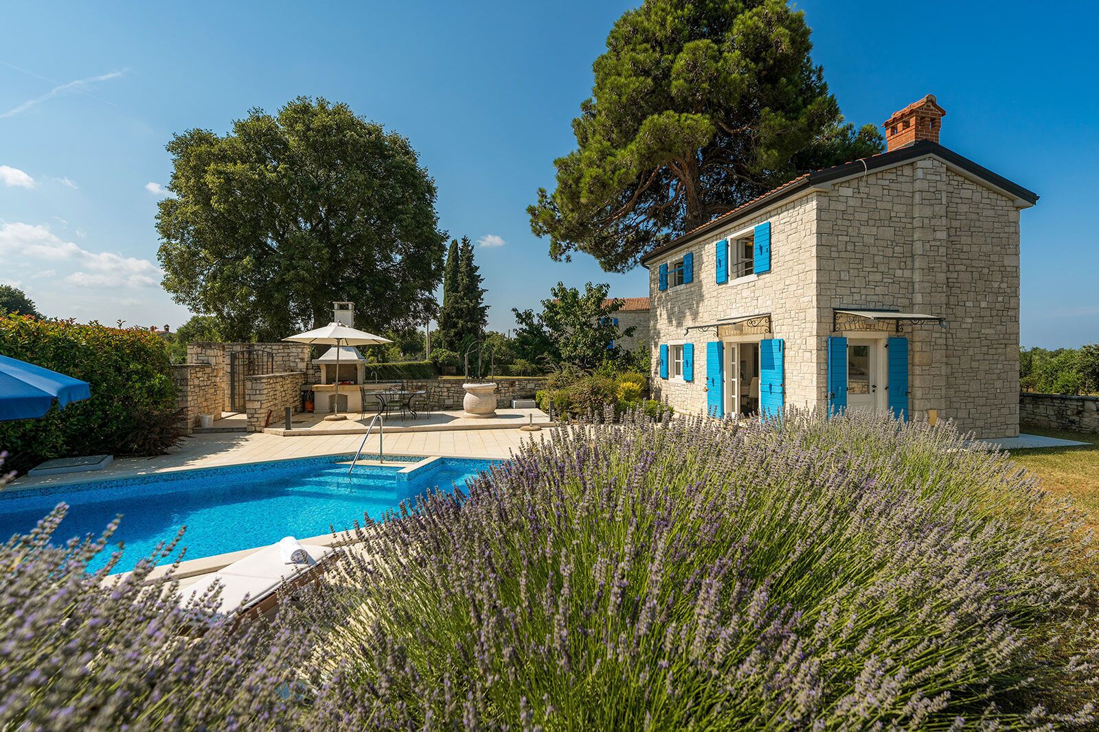 A stone building with blue shutters, a swimming pool, and a garden with lavender.
