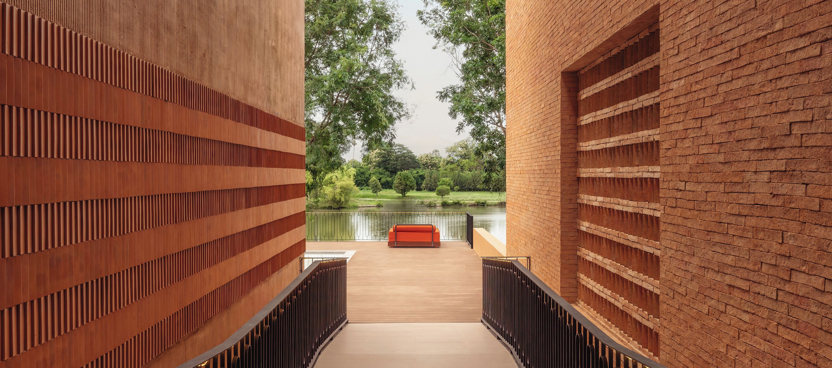 Un pasillo con piso de madera que conduce a una terraza de madera con vista a una masa de agua, con un banco rojo y barandillas de metal.