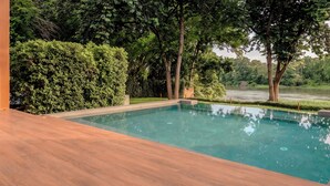 A rectangular swimming pool with a wooden deck area, surrounded by dense greenery and trees, with a body of water in the background. - Hykyo River Kwai Hotel (Kanchanaburi)
