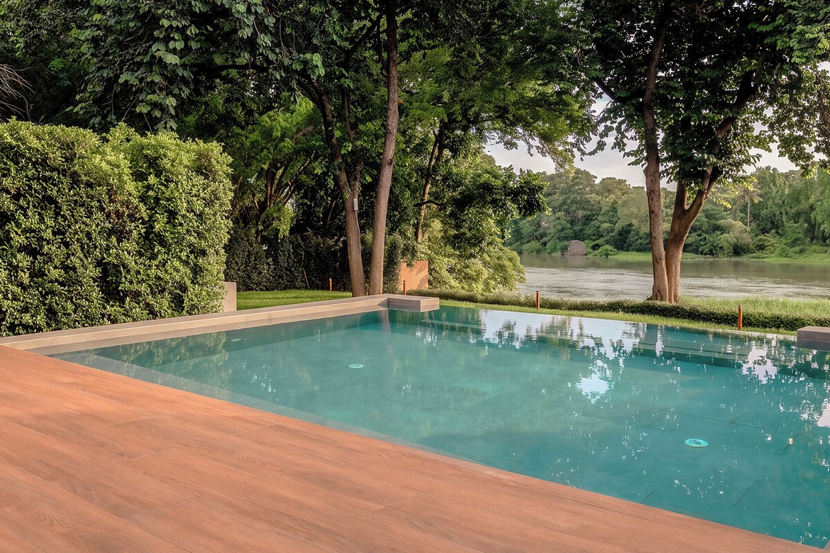 A rectangular swimming pool with a wooden deck area, surrounded by dense greenery and trees, with a body of water in the background.