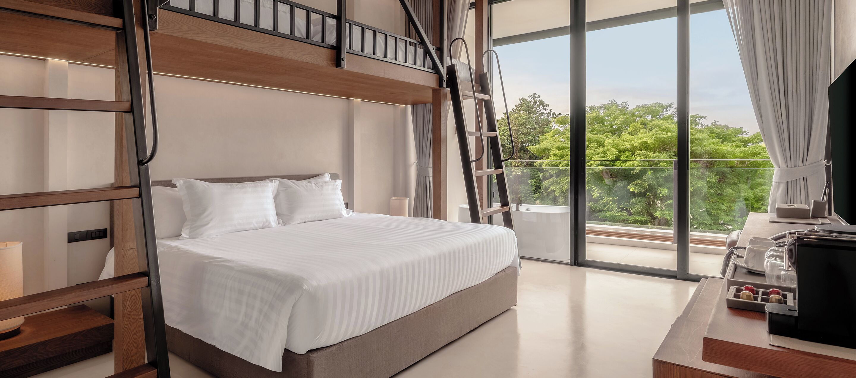 A modern hotel room with a bunk bed, a large window with a view of greenery, and a desk with a laptop and a coffee maker.