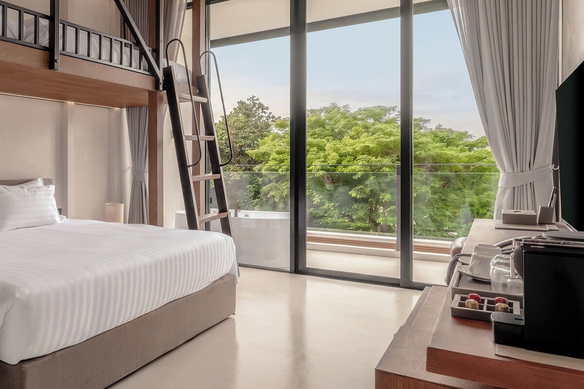 A modern hotel room with a bunk bed, a large window with a view of greenery, and a desk with a laptop and a coffee maker.