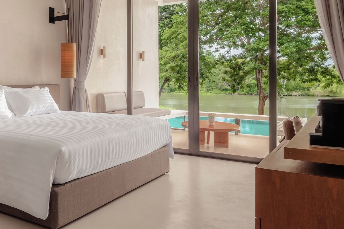 A hotel room with a bed, bedside tables, a TV, and a view of a swimming pool and greenery.