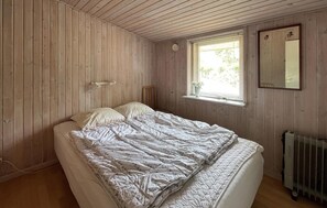 Ein Schlafzimmer mit einer holzverkleideten Wand, einem Einzelbett mit einer Tagesdecke, einem Fenster mit Jalousien, einem Heizkörper und einem Spiegel.