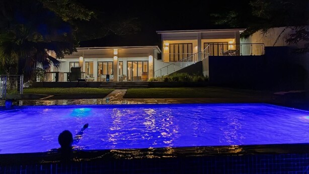 Una piscina con una persona nadando, rodeada por una casa que tiene un porche cubierto y una escalera que lleva a la entrada.