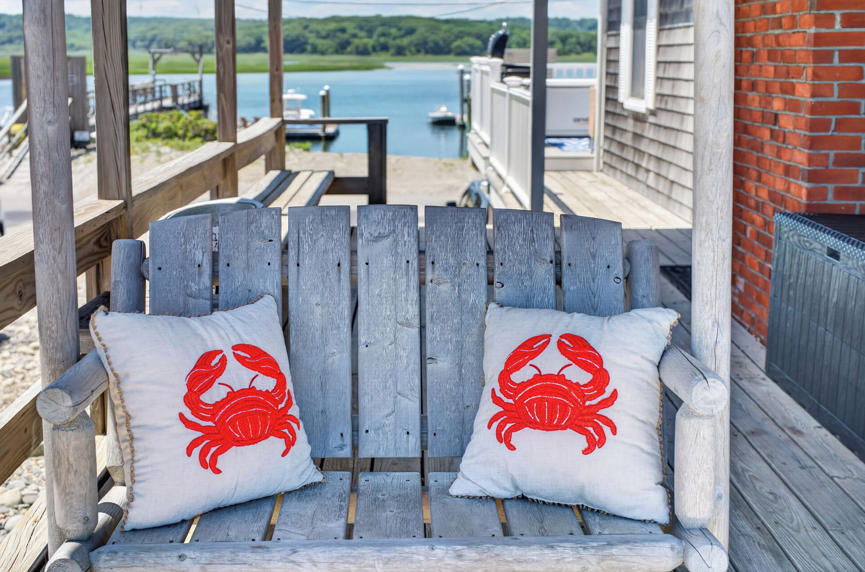 A wooden bench with two white cushions featuring red crab designs, overlooking a body of water with boats and a dock.