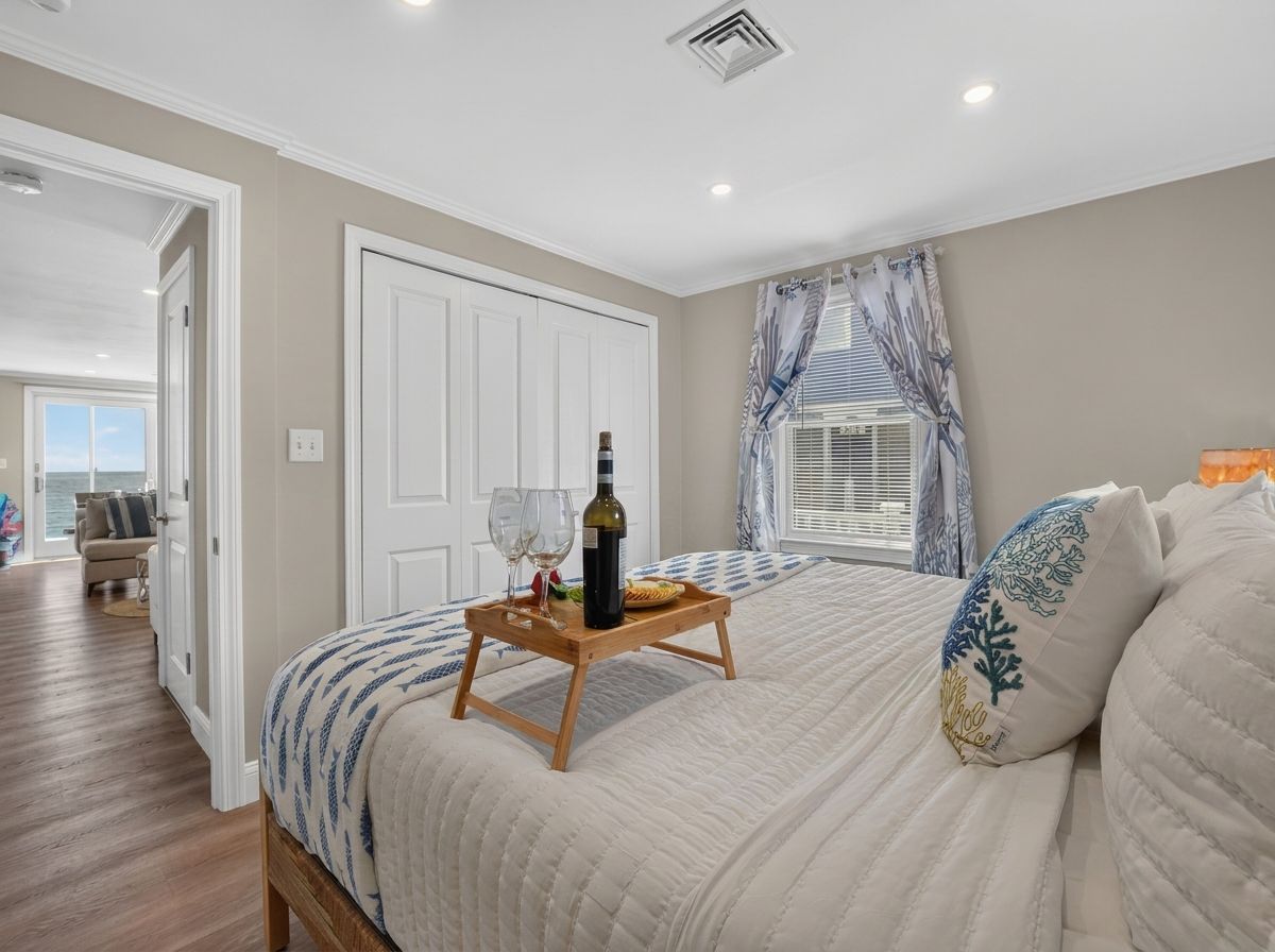 Une chambre à coucher avec un lit, une table de chevet sur laquelle se trouve un plateau avec une bouteille et des verres, une fenêtre avec des rideaux et une porte donnant sur une autre pièce.