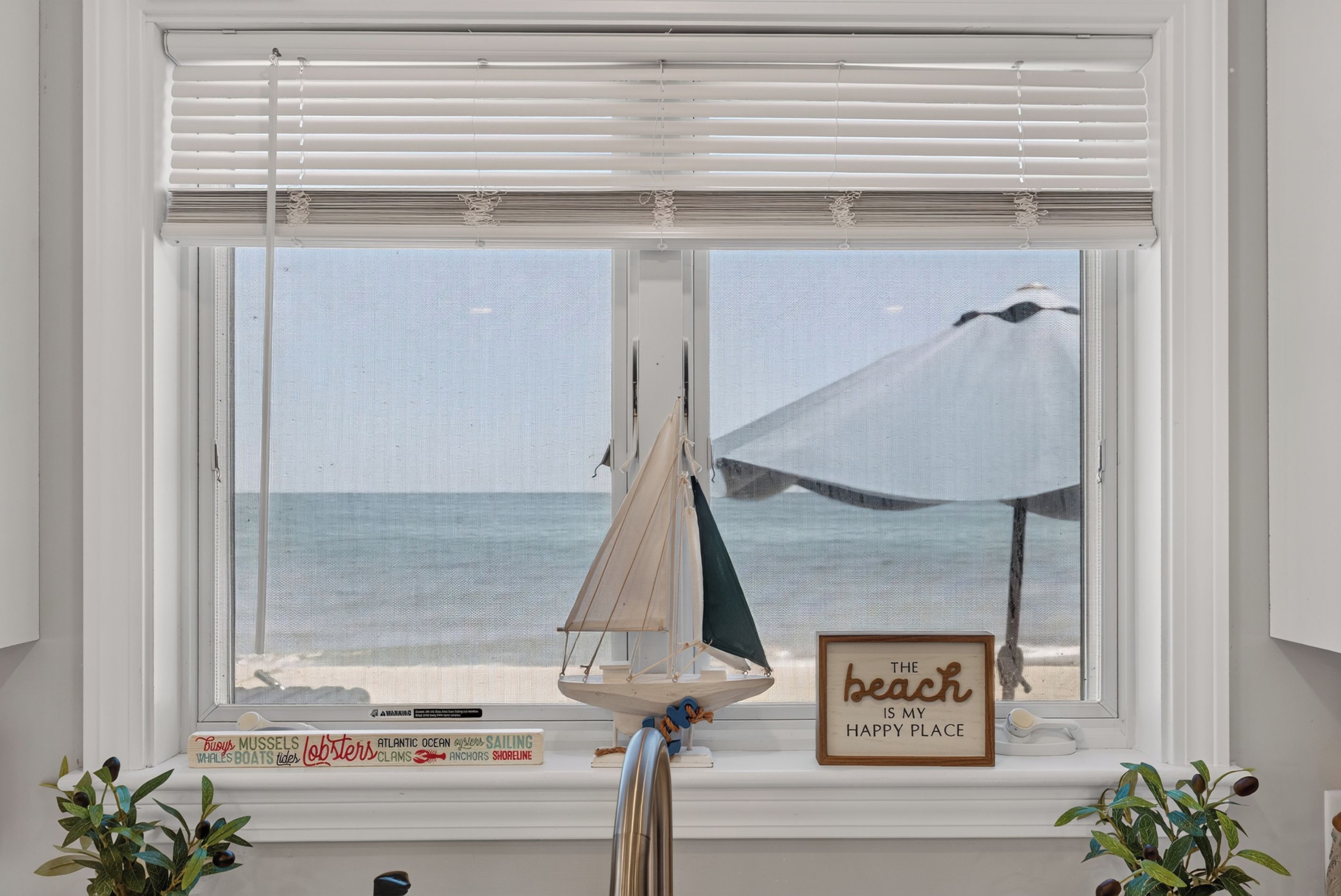 A window with a view of the ocean, a small sailboat model, and a sign that reads 'The beach is my happy place.'