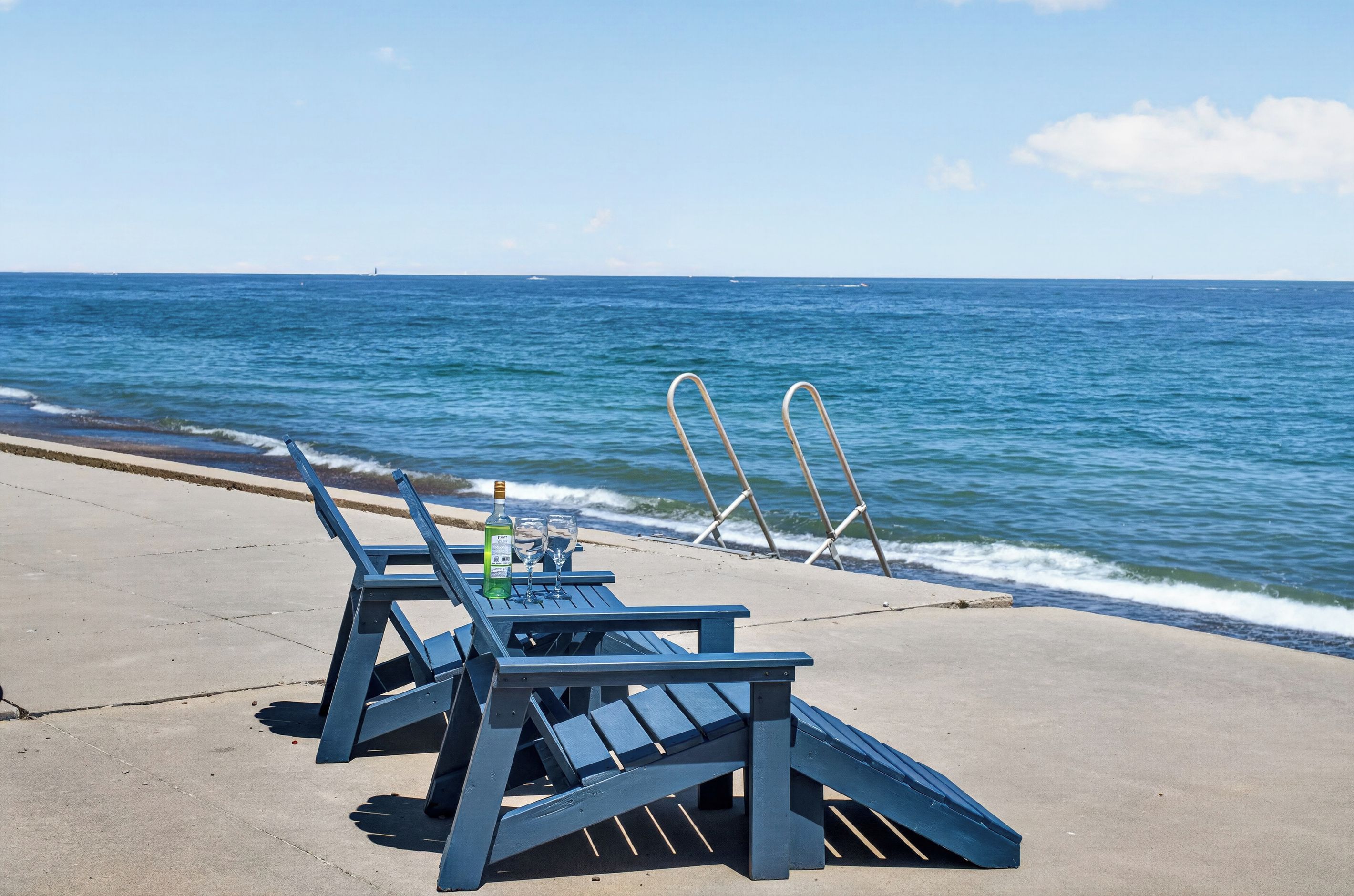 A beachside view with two lounge chairs and a bottle of liquor on a concrete surface.