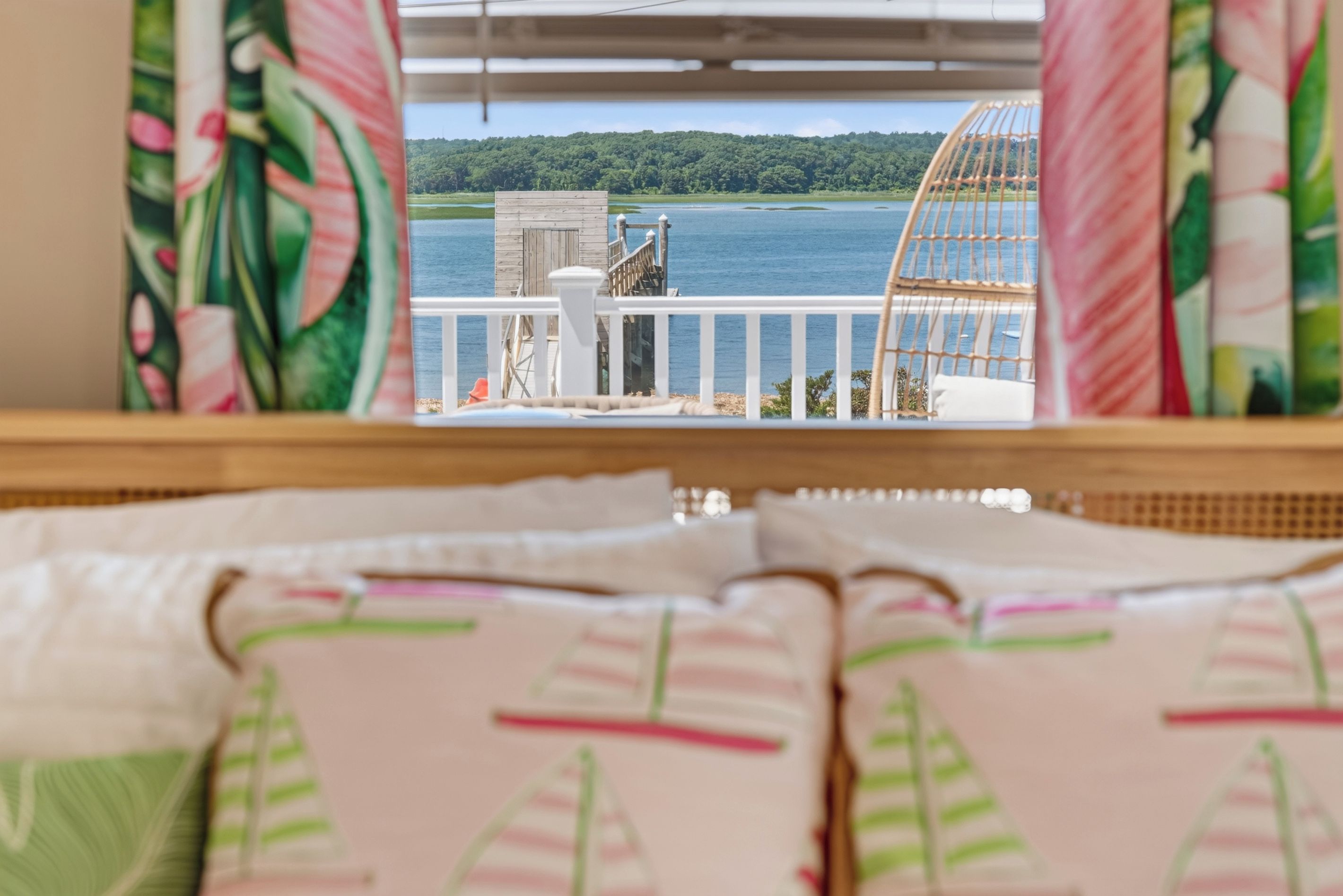 Blick aus einem Fenster auf ein Holzdeck, Geländer in Weiß sowie einen See und einen Wald.