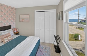 A bedroom with a bed, a window with a view of the sea, a closet, and a wall with a beach-themed wallpaper.