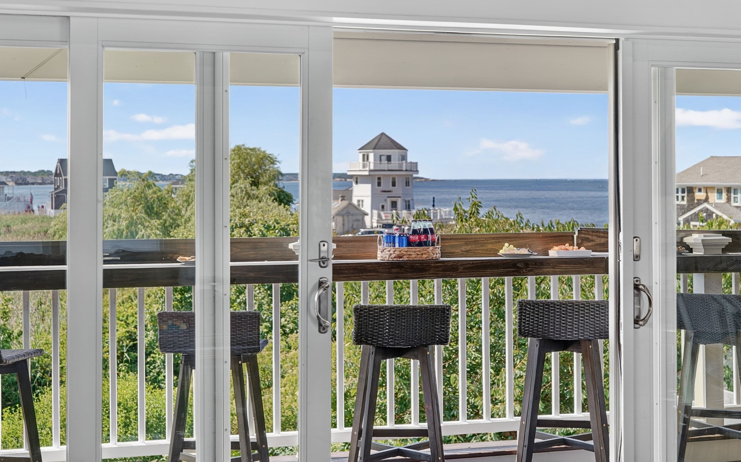 A balcony with wicker chairs and a table, overlooking a coastal view with buildings and greenery.