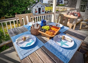 An outdoor dining area with a wooden table set for two, featuring a fruit platter, cheese, and crackers.