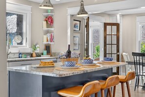 Una cocina con una isla grande, taburetes de bar, iluminación colgante y vistas al exterior.