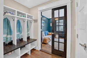 Ein Flur, der zu einem Schlafzimmer mit einer dunkelholzigen Tür, einem weißen Konsolentisch mit Schuhen und einem Wäschebereich mit Handtüchern und Kleiderbügeln führt.