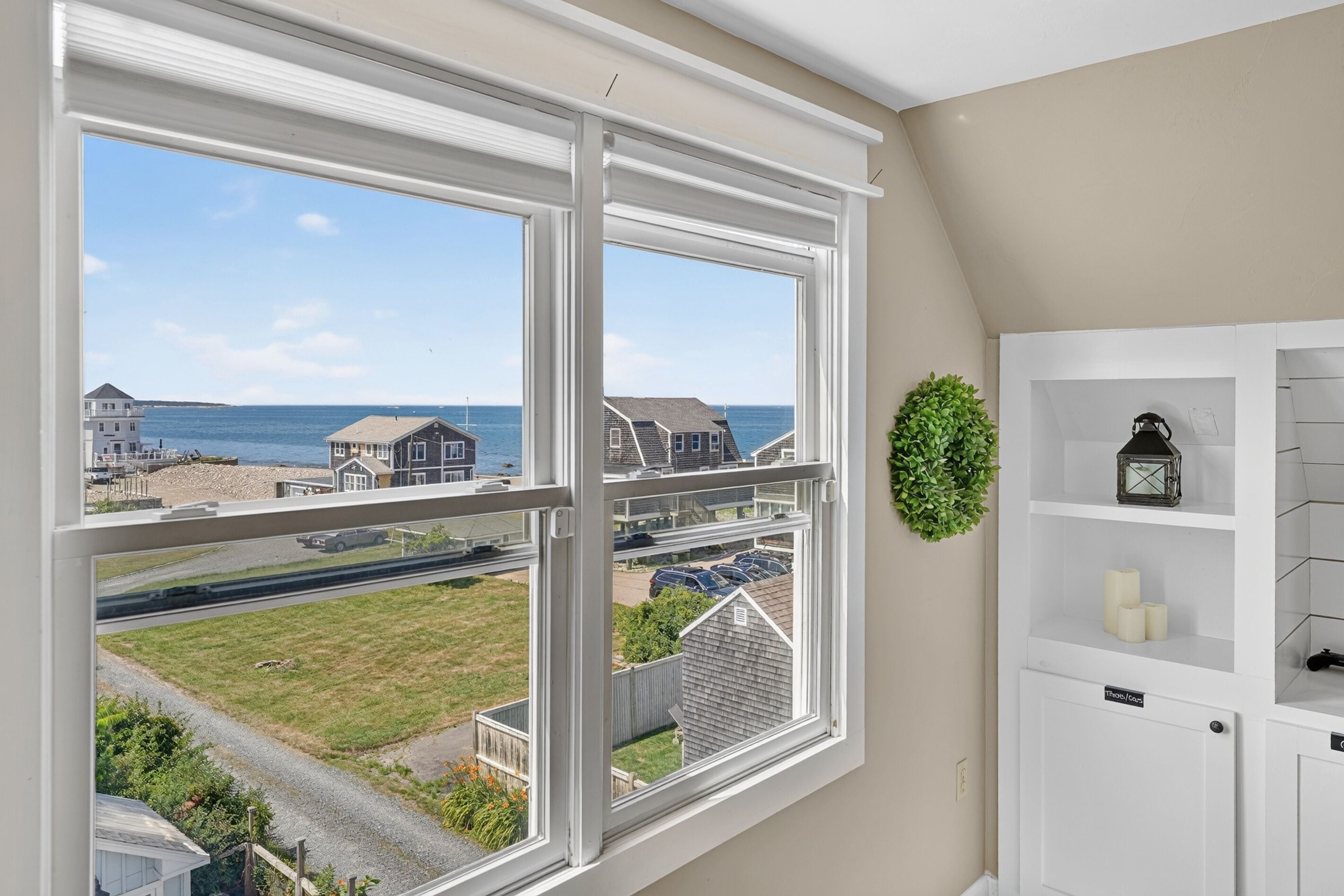 A room with a large window offering a view of the sea and neighboring houses.