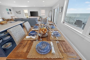 A dining area with a wooden table set for a meal, surrounded by wicker furniture and a view of the sea.