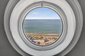 Vista per un finestral circular d'una platja amb roques i un cel clar.