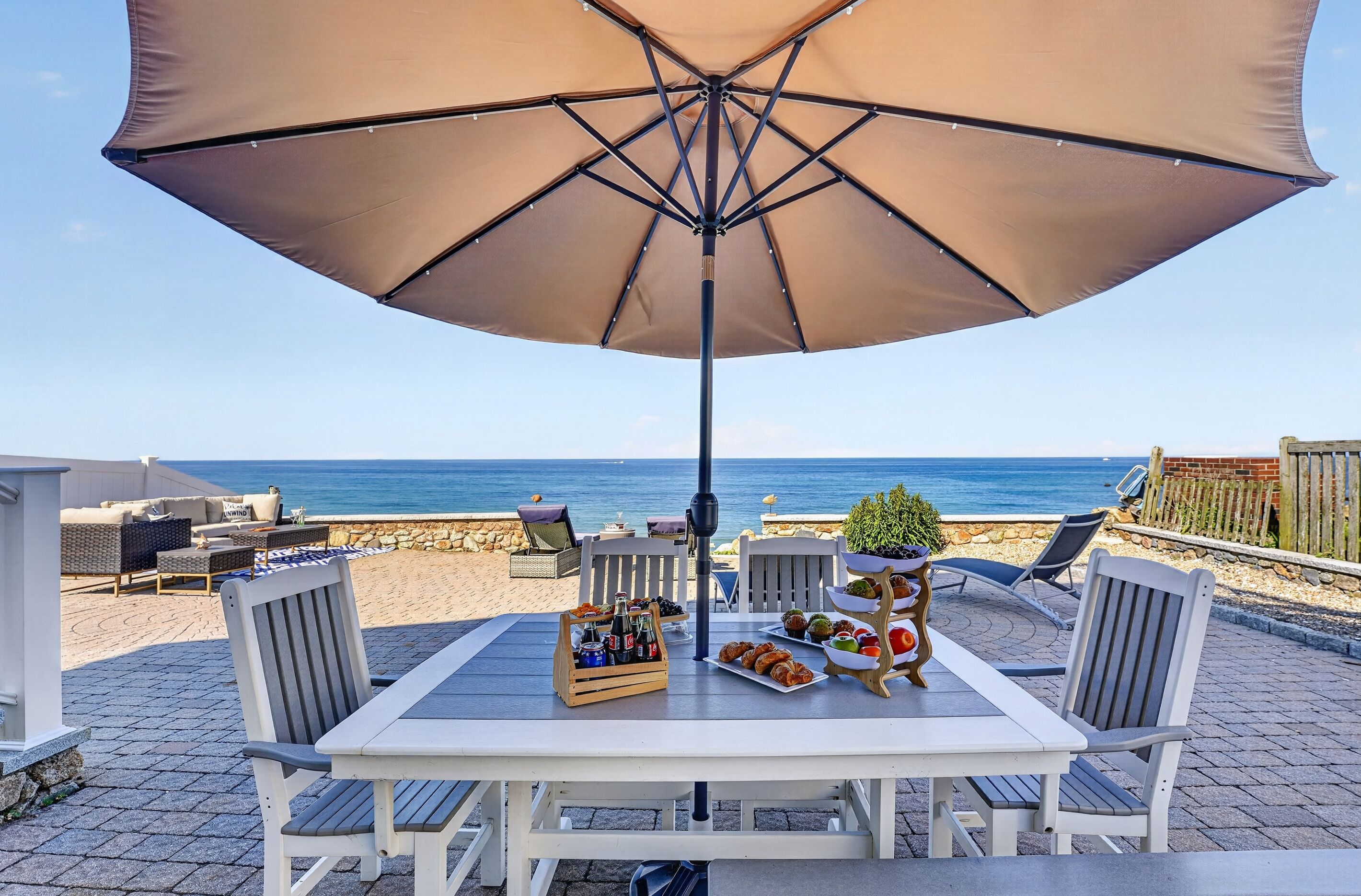 A table with a large umbrella, surrounded by chairs, with a view of the sea.