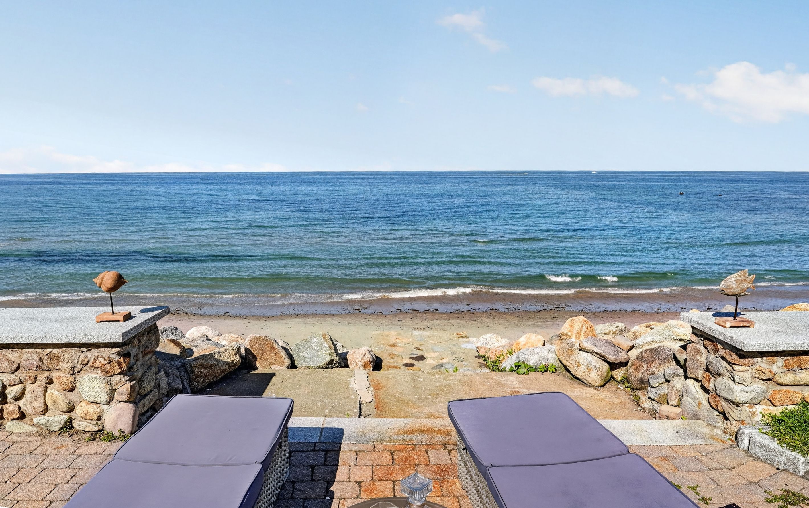 Un espace de détente aménagé au bord de la plage, avec deux chaises longues et des sculptures d’oiseaux décoratives.